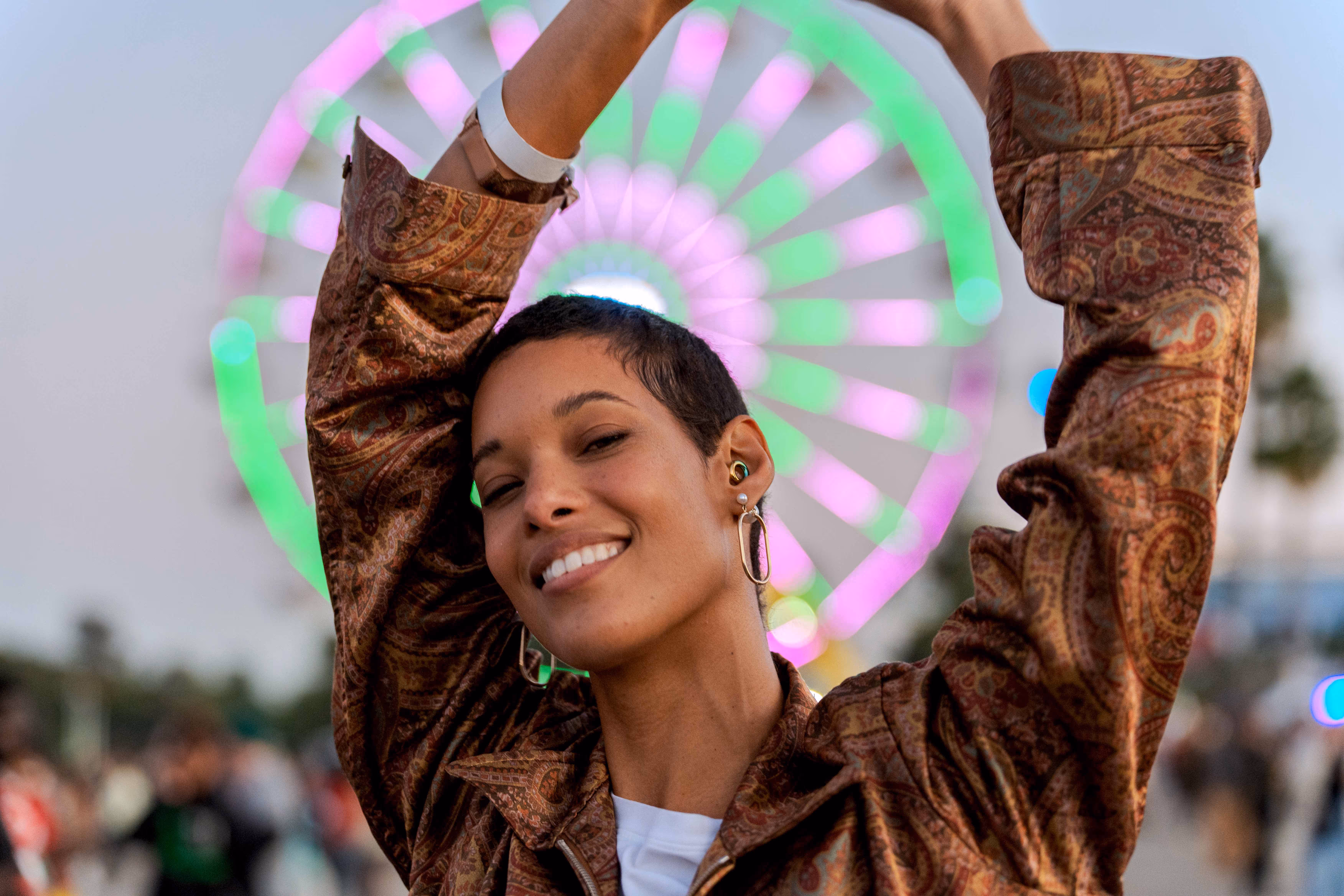 Smiling woman wearing earplugs at an outdoor festival with a brightly lit ferris wheel in the background, emphasizing safe listening at loud events.