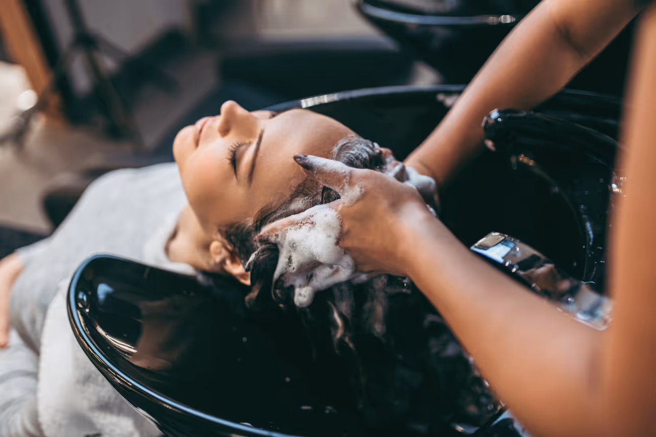A woman reclines at a salon sink with her eyes closed while a stylist gently shampoos her hair. The scene conveys calmness and relaxation in a typical hair salon setting.