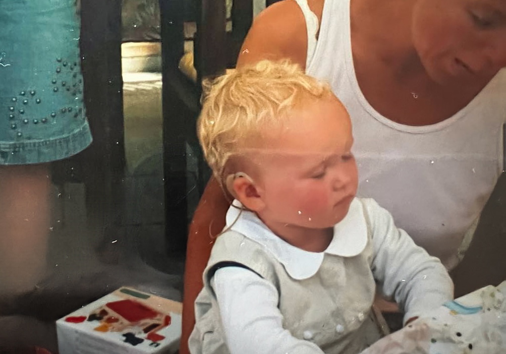 A close-up of a baby with light curly hair sitting on a woman’s lap, looking downward. A cochlear implant is visible behind the baby’s ear.