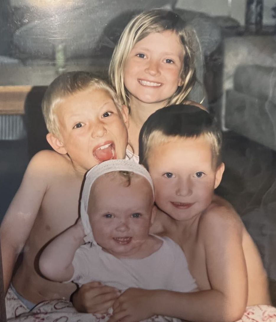 Four smiling children cuddle together on a couch. The youngest, a baby wearing a bonnet, is seated in front and grinning while the others gather close behind.