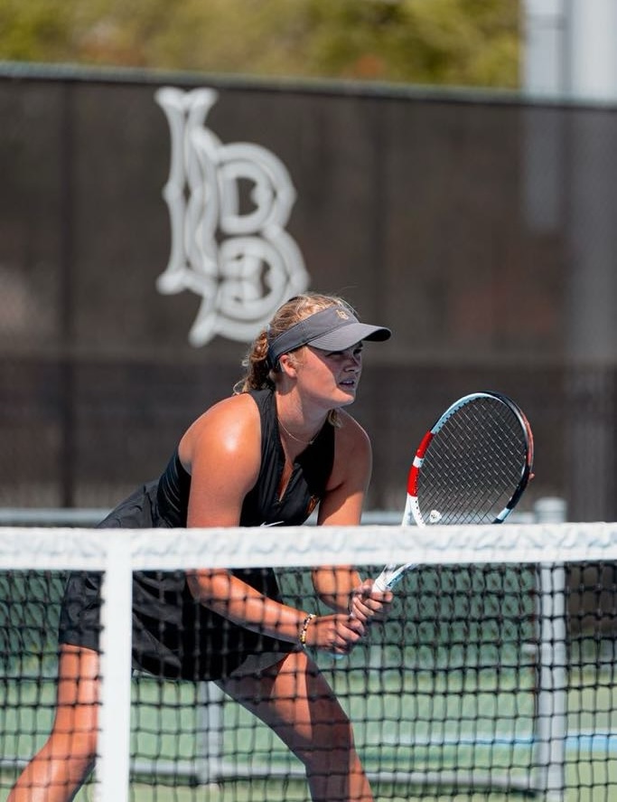 Mathilde Tranberg, a cochlear implant advocate and former Division I tennis player, stands focused at the net during a tennis match at Long Beach State. She wears a black athletic dress and visor, holding her racket ready in both hands.