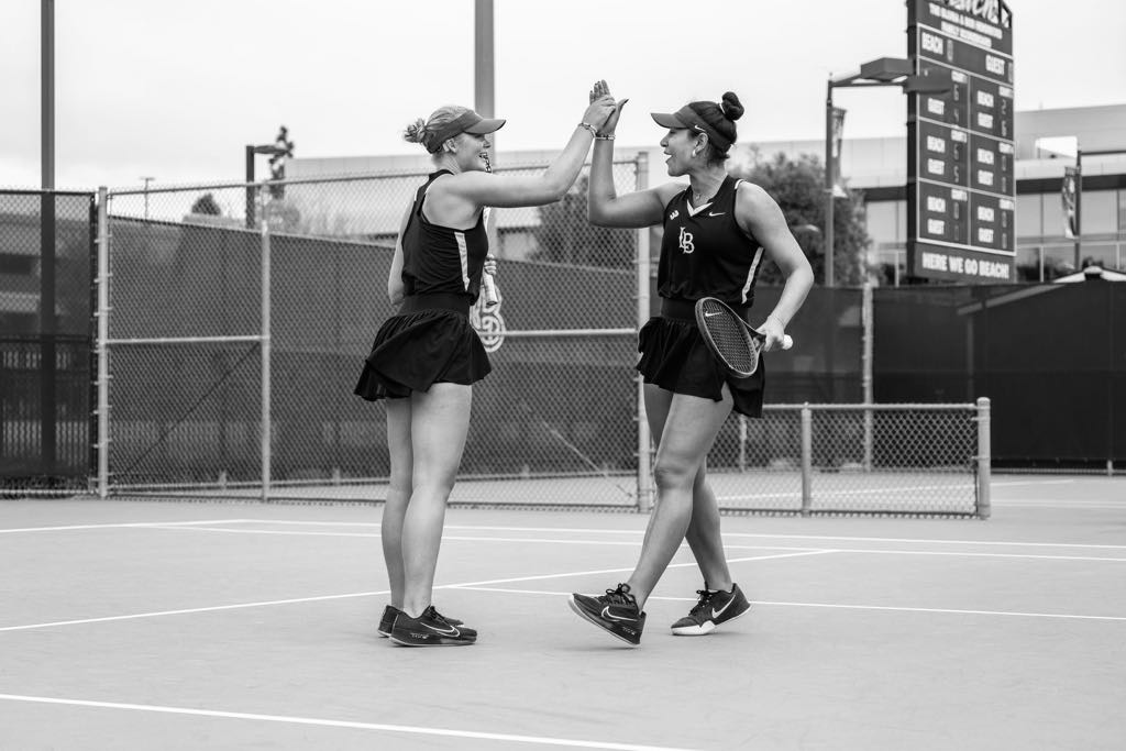 Black-and-white photo of two female tennis players, Mathilde Tranberg (left) and her doubles partner, on an outdoor court at Long Beach State. Mathilde, a young white woman with a blonde bun and sun visor, wears a sleeveless athletic top and tennis skirt as she smiles and high-fives her teammate mid-match. Both players hold rackets, and a scoreboard and fencing are visible in the background.