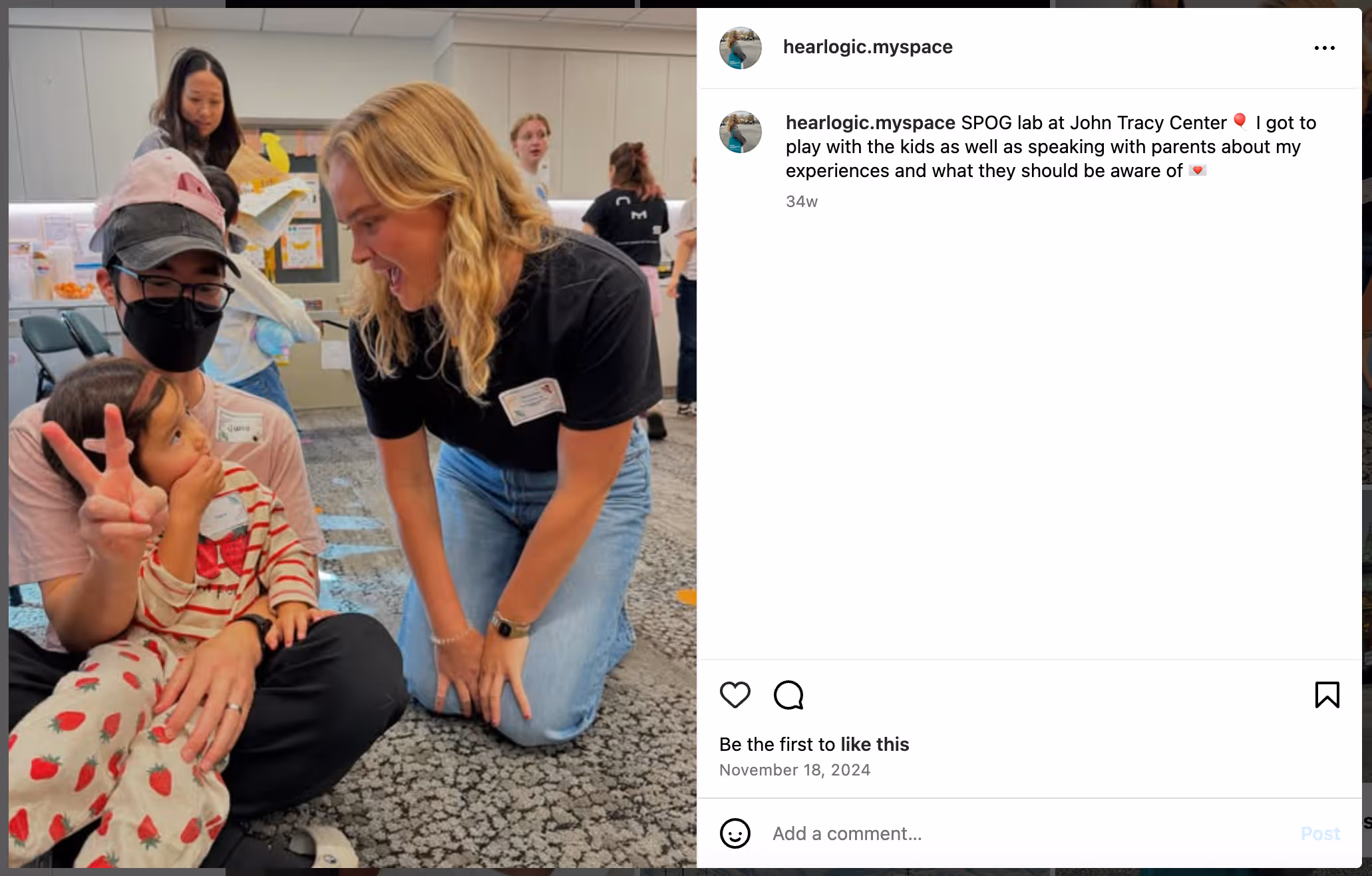 Mathilde Tranberg, a young blonde woman with light skin, kneels and smiles at a child sitting on a parent’s lap at the John Tracy Center. She’s wearing a black shirt and jeans during a community outreach event about hearing loss.