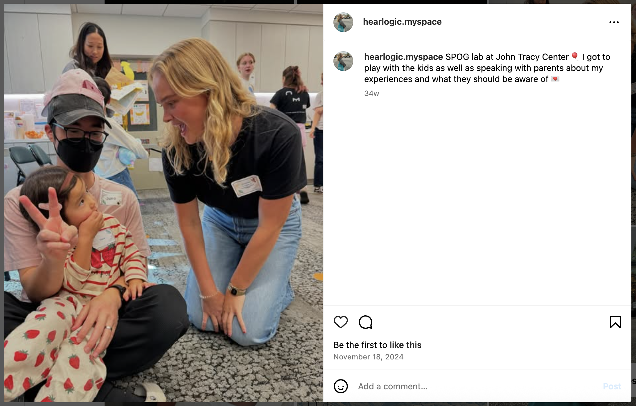 Mathilde Tranberg, a young blonde woman with light skin, kneels and smiles at a child sitting on a parent’s lap at the John Tracy Center. She’s wearing a black shirt and jeans during a community outreach event about hearing loss.