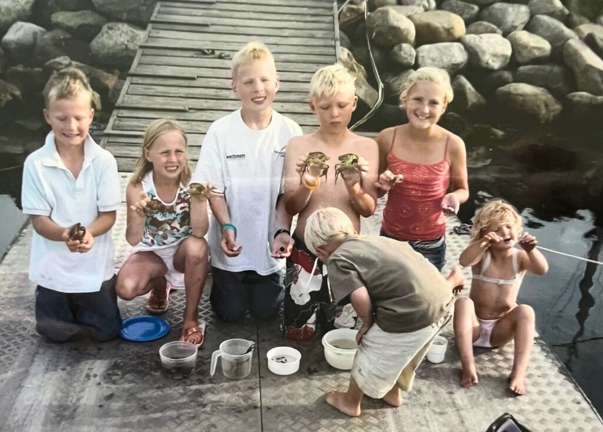 A group of children kneel and stand on a dock, proudly holding crabs and fishing gear. Behind them, a small wooden ramp leads to a rocky shoreline.