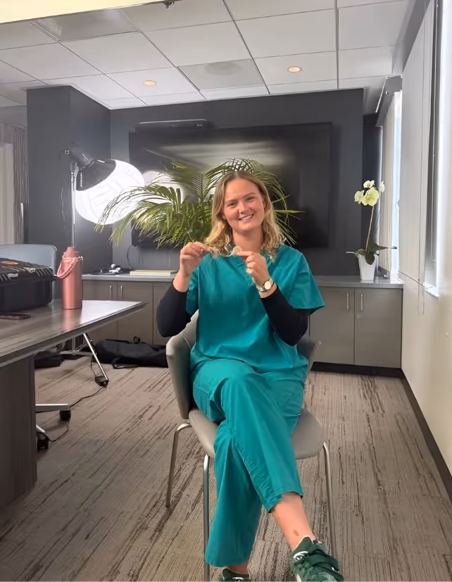 Mathilde Tranberg, a young white woman with long, wavy blonde hair, sits smiling in green scrubs at Pacific Neuroscience Institute. She holds a cochlear implant device in her hands, seated in a modern office with a large monitor and studio lights in the background.