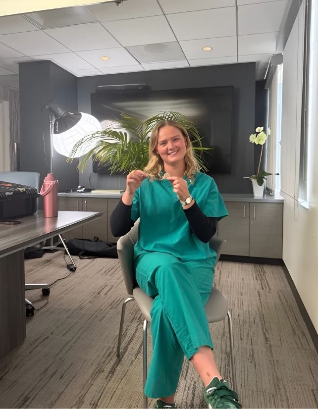 Mathilde Tranberg, a young white woman with long, wavy blonde hair, sits smiling in green scrubs at Pacific Neuroscience Institute. She holds a cochlear implant device in her hands, seated in a modern office with a large monitor and studio lights in the background.