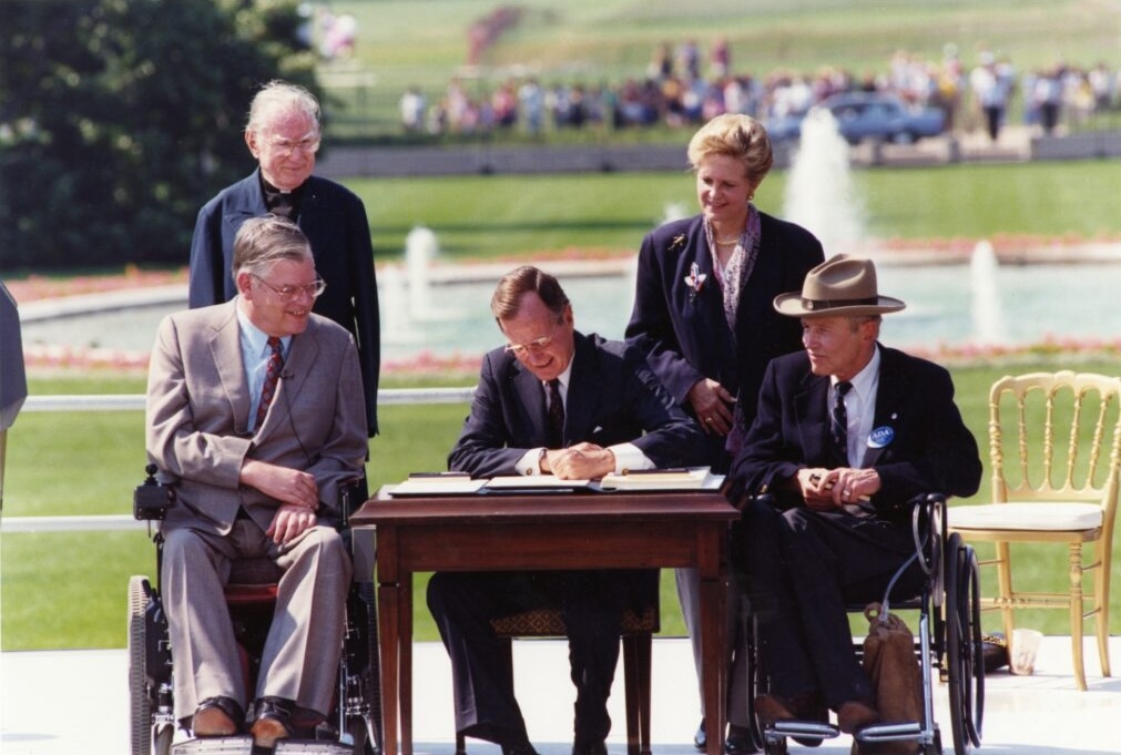 President George H. W. Bush sits at a wooden desk outdoors, signing the Americans with Disabilities Act (ADA) into law on July 26, 1990. He is surrounded by key disability rights advocates, including two men in wheelchairs. Behind them, a large fountain and the White House lawn are visible, with onlookers gathered in the background. The moment marks a historic milestone in civil rights legislation for people with disabilities.