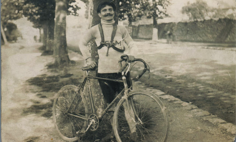 Black-and-white photo of Eugène Rubens-Alcais, founder of the Deaflympics, standing beside a vintage bicycle and smiling. He wears a cycling outfit marked with “F.S.S.M.” and a scarf around his neck.