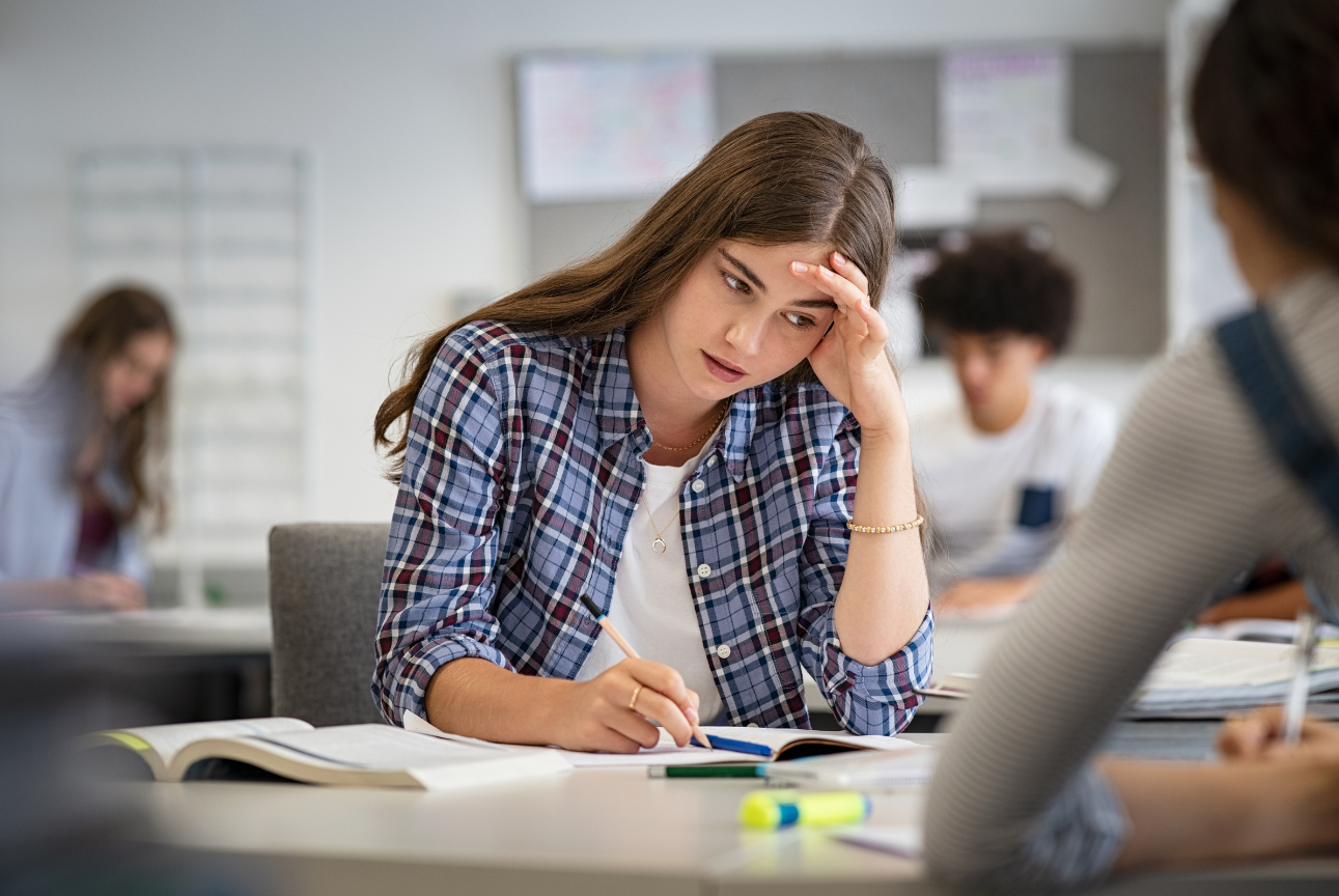 A college student sits at a classroom desk, leaning on one hand while writing in a notebook. She appears focused and slightly stressed, surrounded by open textbooks and highlighters. Other students are blurred in the background.