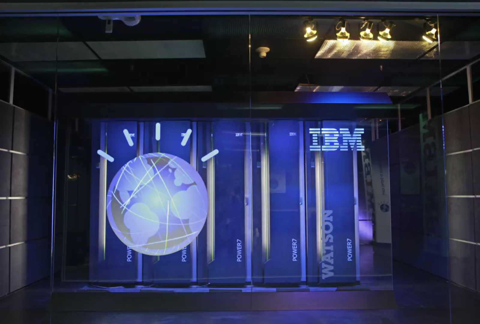The illuminated IBM Watson supercomputer display shows the recognizable blue globe logo with network lines, alongside servers labeled “Power7” inside a glass-walled exhibit.