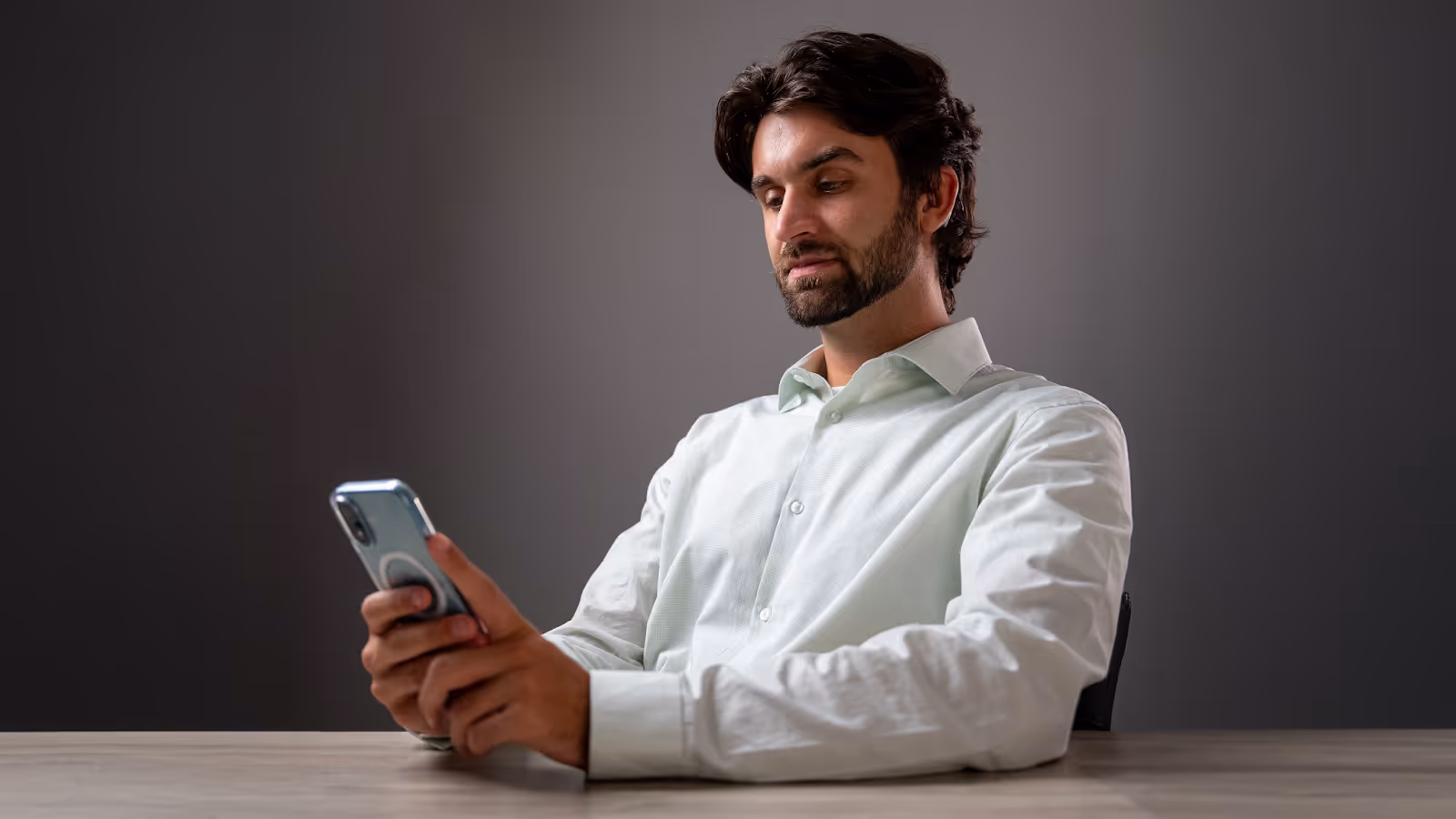 A man with dark hair and a trimmed beard sits at a desk, looking at his smartphone with a slight smile. He wears a light green button-down shirt against a neutral gray background.