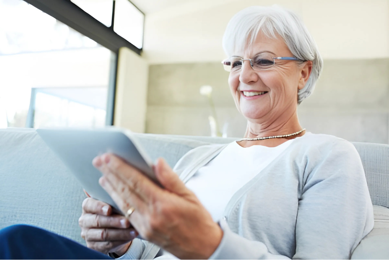An older woman with short white hair and glasses sits comfortably on a couch, smiling as she uses a tablet. Natural light fills the modern living room, highlighting her engagement with the device.