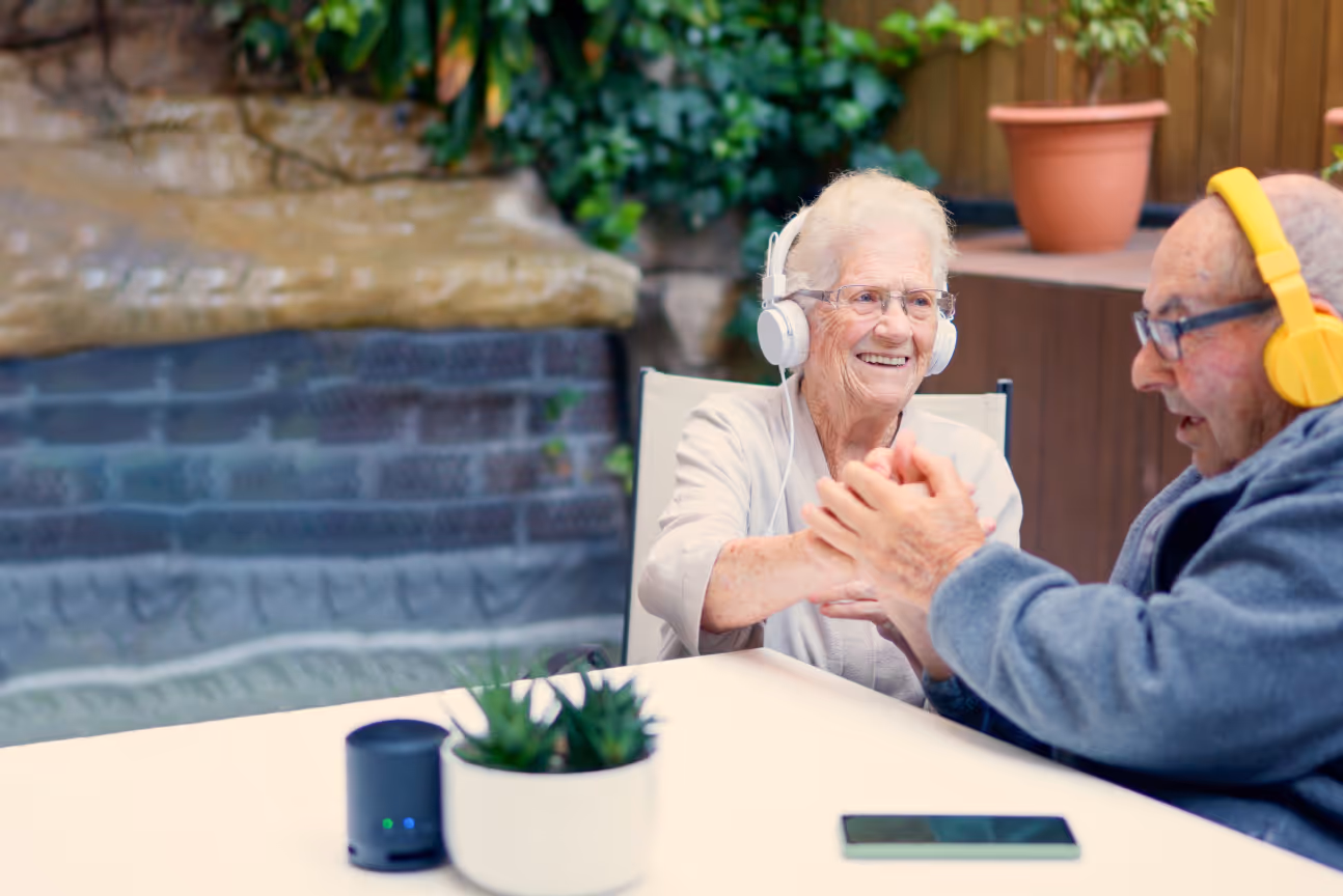 An older woman and man sit together outdoors, both wearing headphones and smiling warmly. The woman clasps the man’s hands across the table, while a small speaker, potted plant, and smartphone rest in front of them.