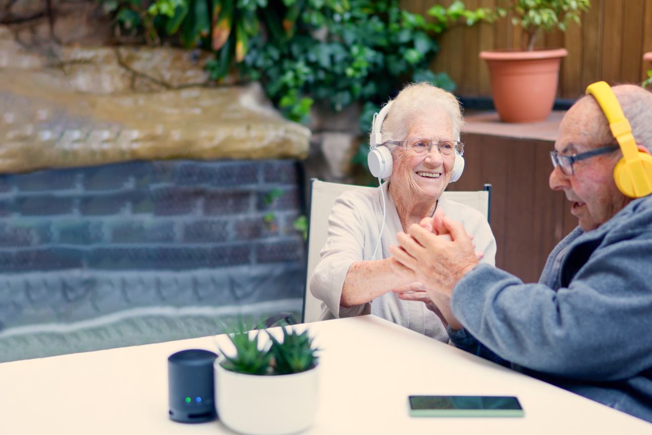 An older woman and man sit together outdoors, both wearing headphones and smiling warmly. The woman clasps the man’s hands across the table, while a small speaker, potted plant, and smartphone rest in front of them.