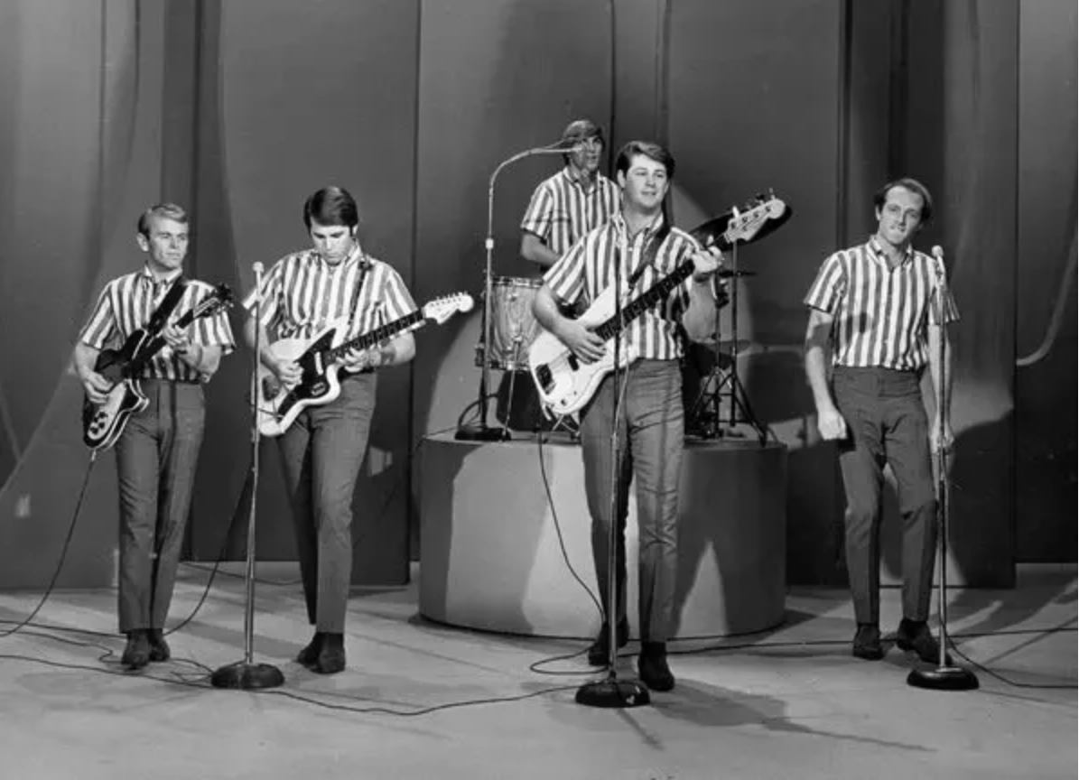 The Beach Boys perform on stage in matching striped shirts, playing guitars and singing into microphones.