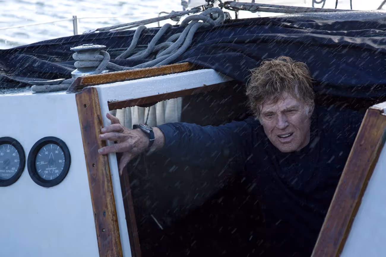 An older man with wet hair emerges from the cabin of a sailboat during heavy rain, gripping the doorway.