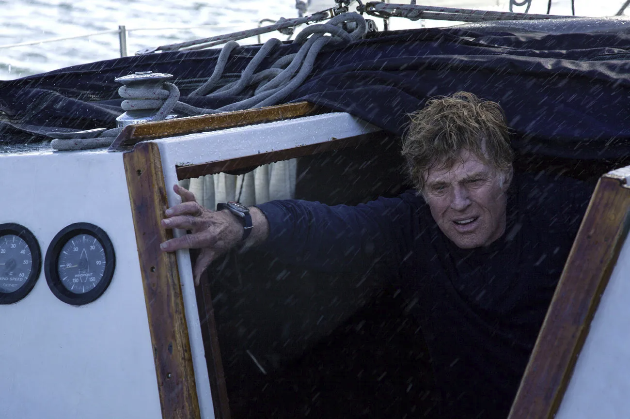 An older man with wet hair emerges from the cabin of a sailboat during heavy rain, gripping the doorway.