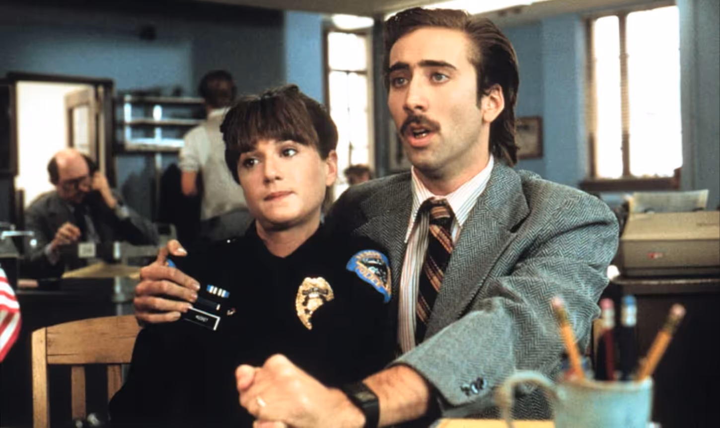 A man in a suit and tie with a mustache holds a woman in a police uniform close while sitting at a desk in a police station.