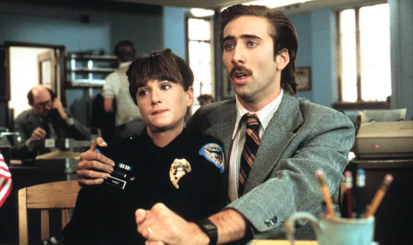 A man in a suit and tie with a mustache holds a woman in a police uniform close while sitting at a desk in a police station.
