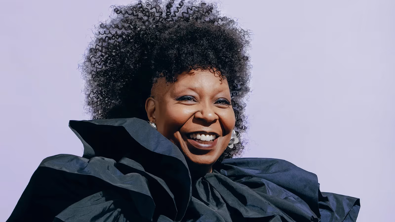 Whoopi Goldberg smiles brightly in a dramatic black ruffled outfit against a pale purple background.