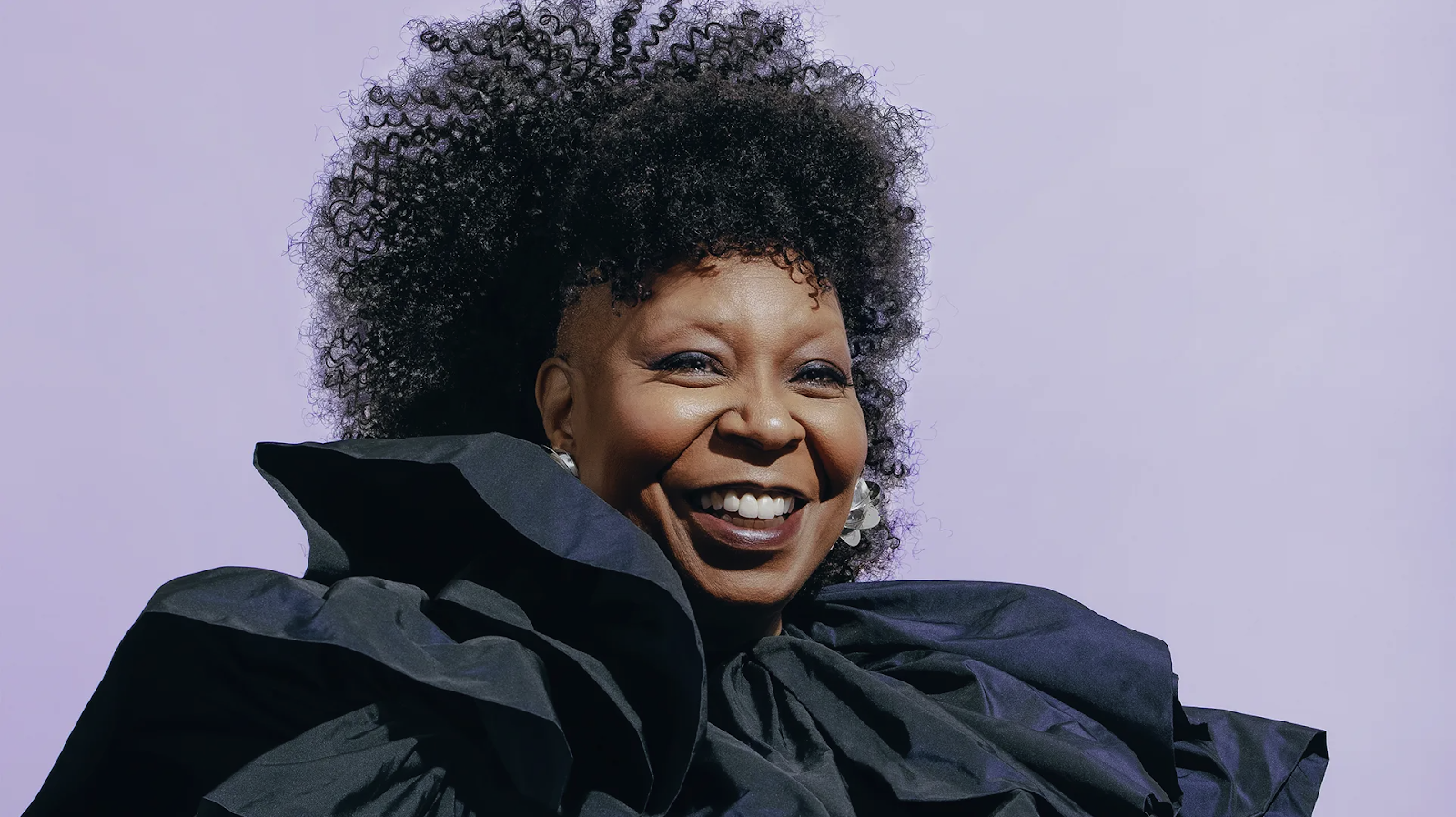 Whoopi Goldberg smiles brightly in a dramatic black ruffled outfit against a pale purple background.