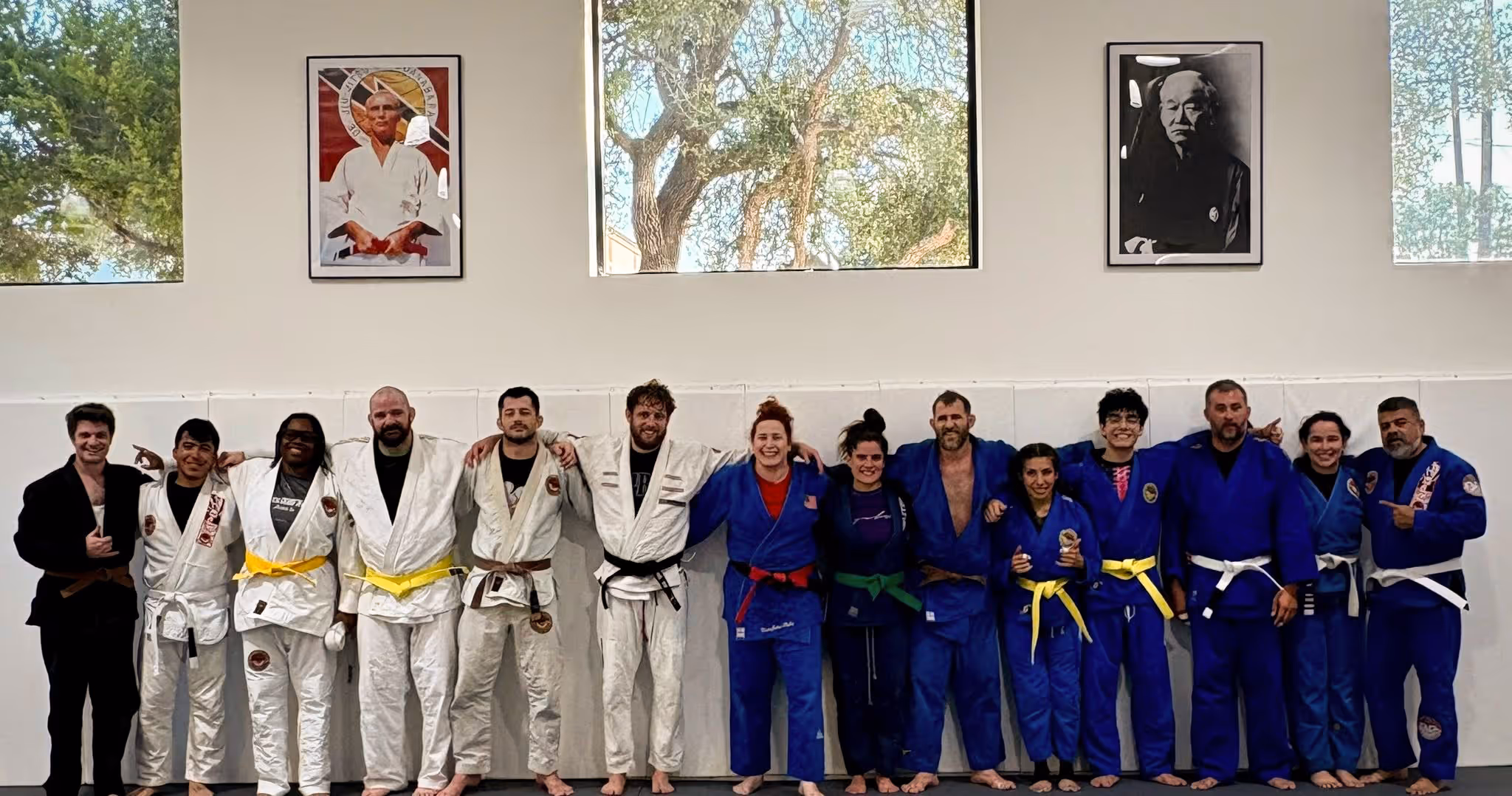 Nina Cutro-Kelly stands at the center of her Judo teammates during a training session at RPBJJ. The group, dressed in white and blue gis with various belt ranks, smiles side by side in a show of unity and camaraderie on the dojo mat.