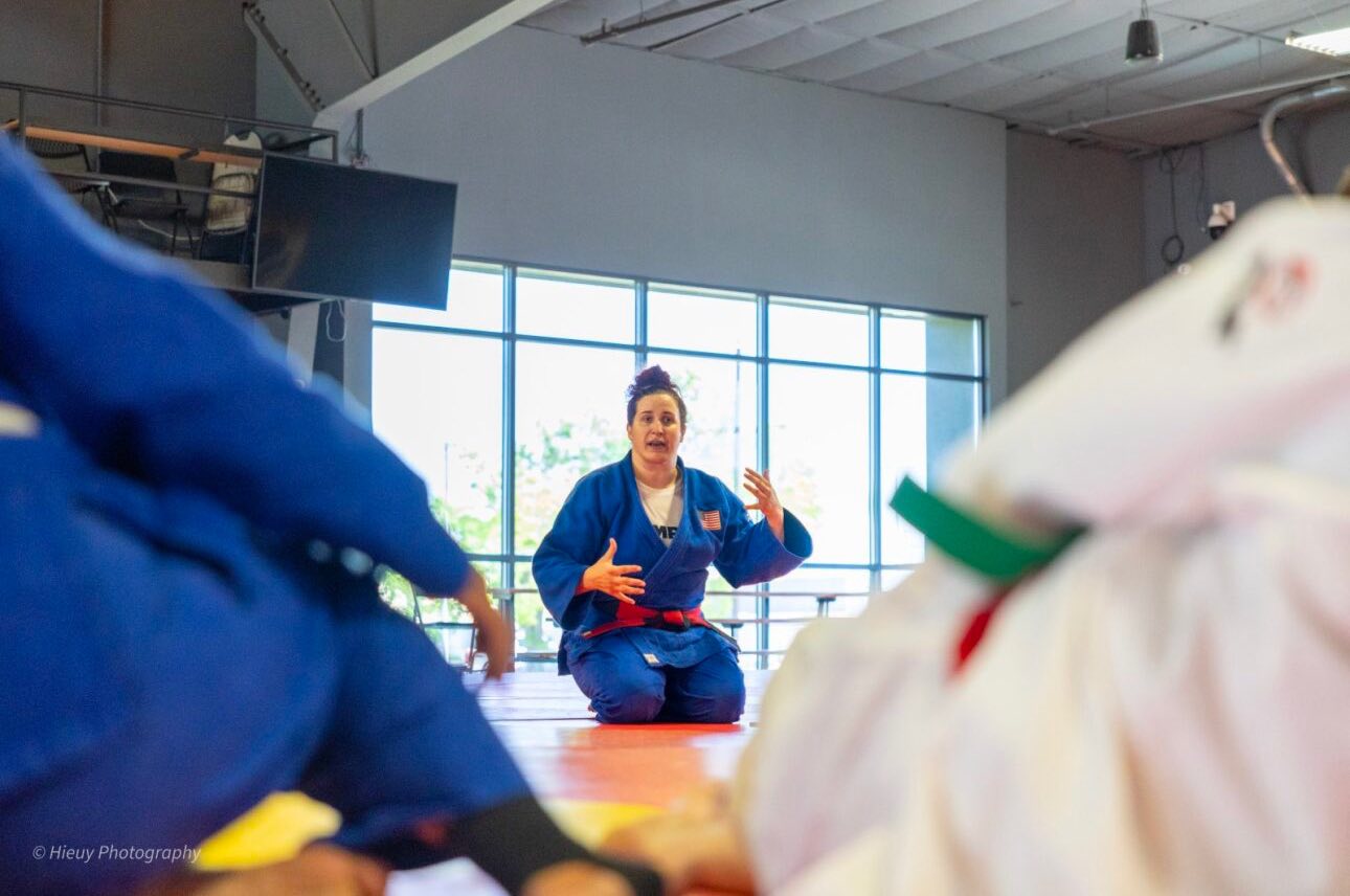 Nina Cutro-Kelly kneels on the mat in her blue gi, gesturing animatedly as she explains Judo techniques to athletes gathered around her.