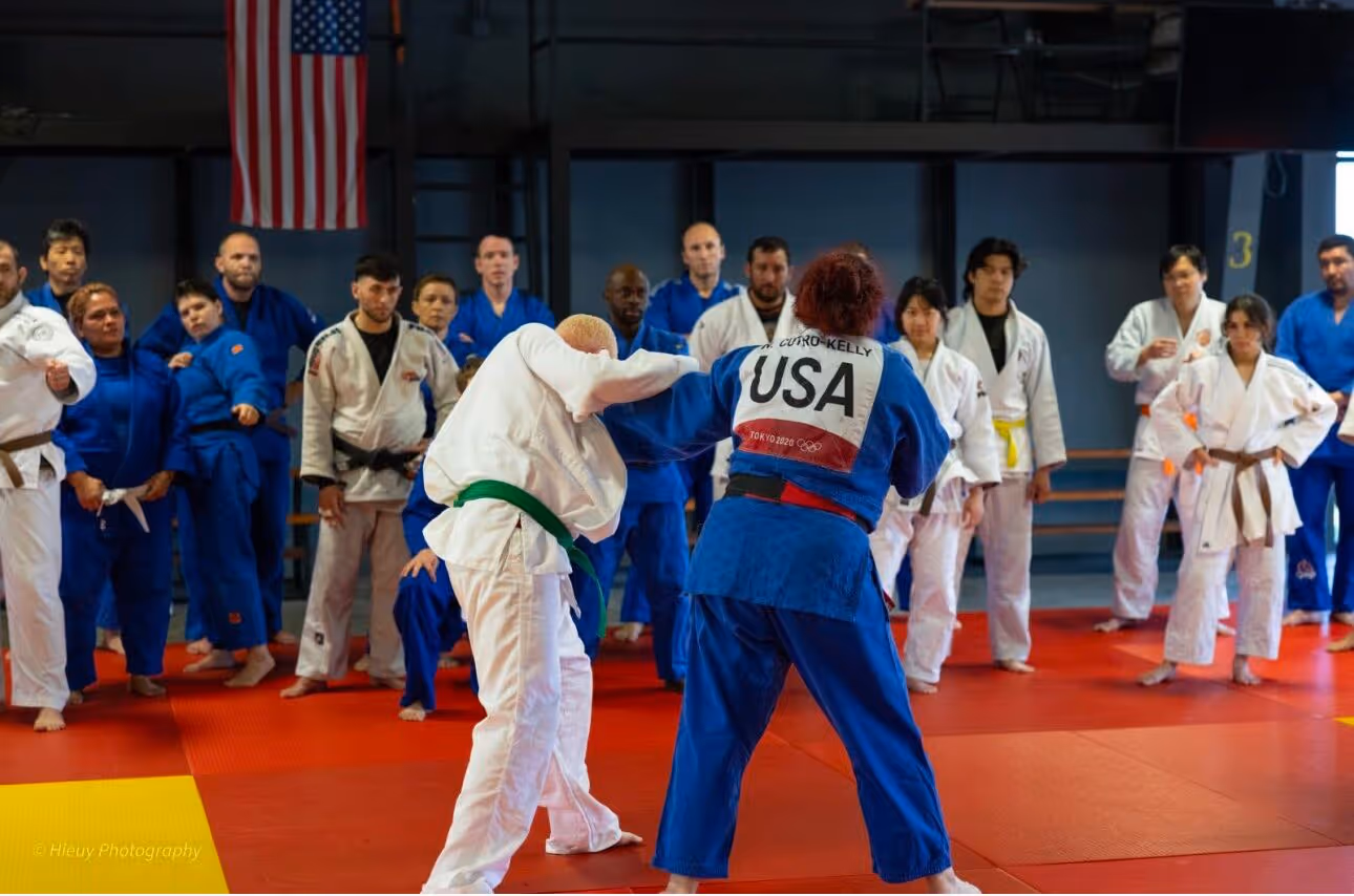 Alt text: Wearing her blue gi with “USA” on the back, Nina Cutro-Kelly demonstrates a Judo throw while athletes in gis watch closely from the edge of the mat beneath an American flag.
