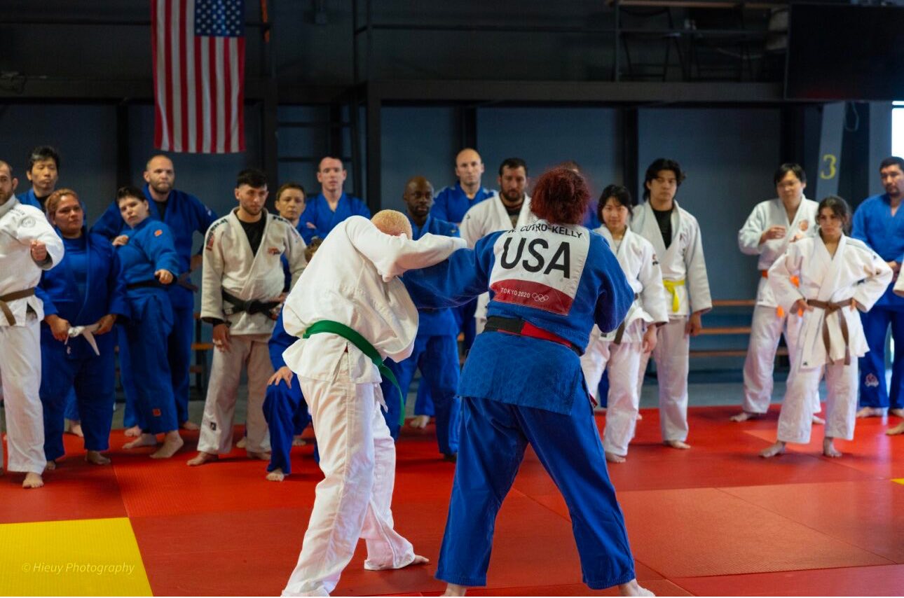 Alt text: Wearing her blue gi with “USA” on the back, Nina Cutro-Kelly demonstrates a Judo throw while athletes in gis watch closely from the edge of the mat beneath an American flag.
