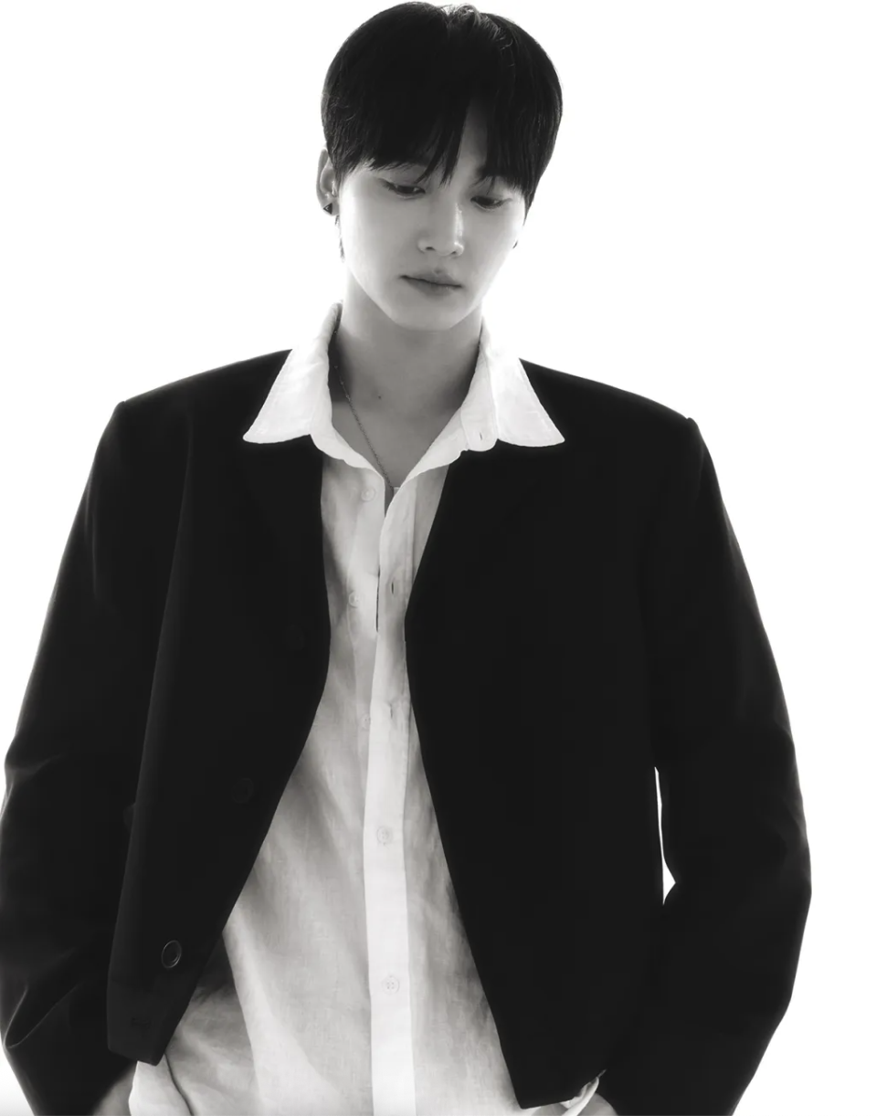 A black-and-white portrait of a Big Ocean member wearing a simple white T-shirt, standing with head slightly bowed and hands behind his back.