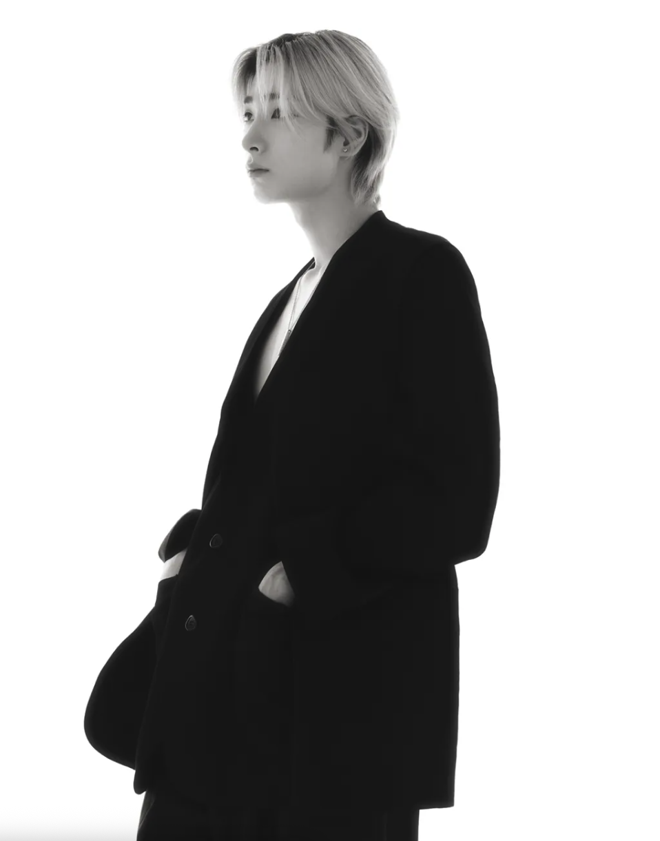 A black-and-white portrait of a Big Ocean member with light hair, wearing a black blazer over a white shirt, gazing to the side with hands in pockets.