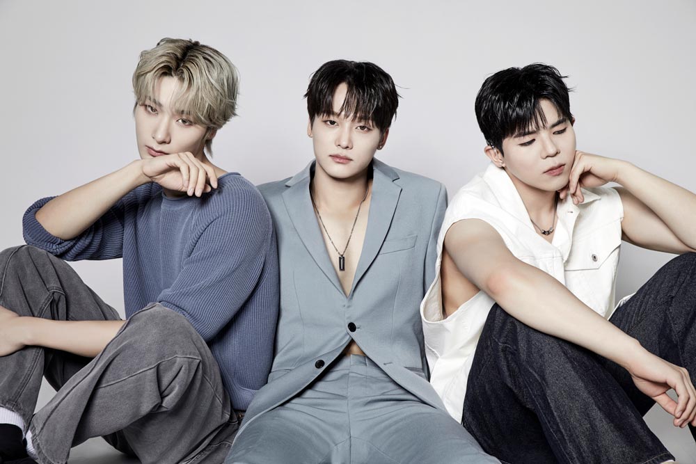 The three members of Big Ocean sit against a light gray background. From left to right: one in a blue sweater with ash-blonde hair, one in a pale blue suit with dark hair, and one in a white sleeveless shirt with dark hair. Their styling is modern and minimal, capturing the group’s cool, confident presence.