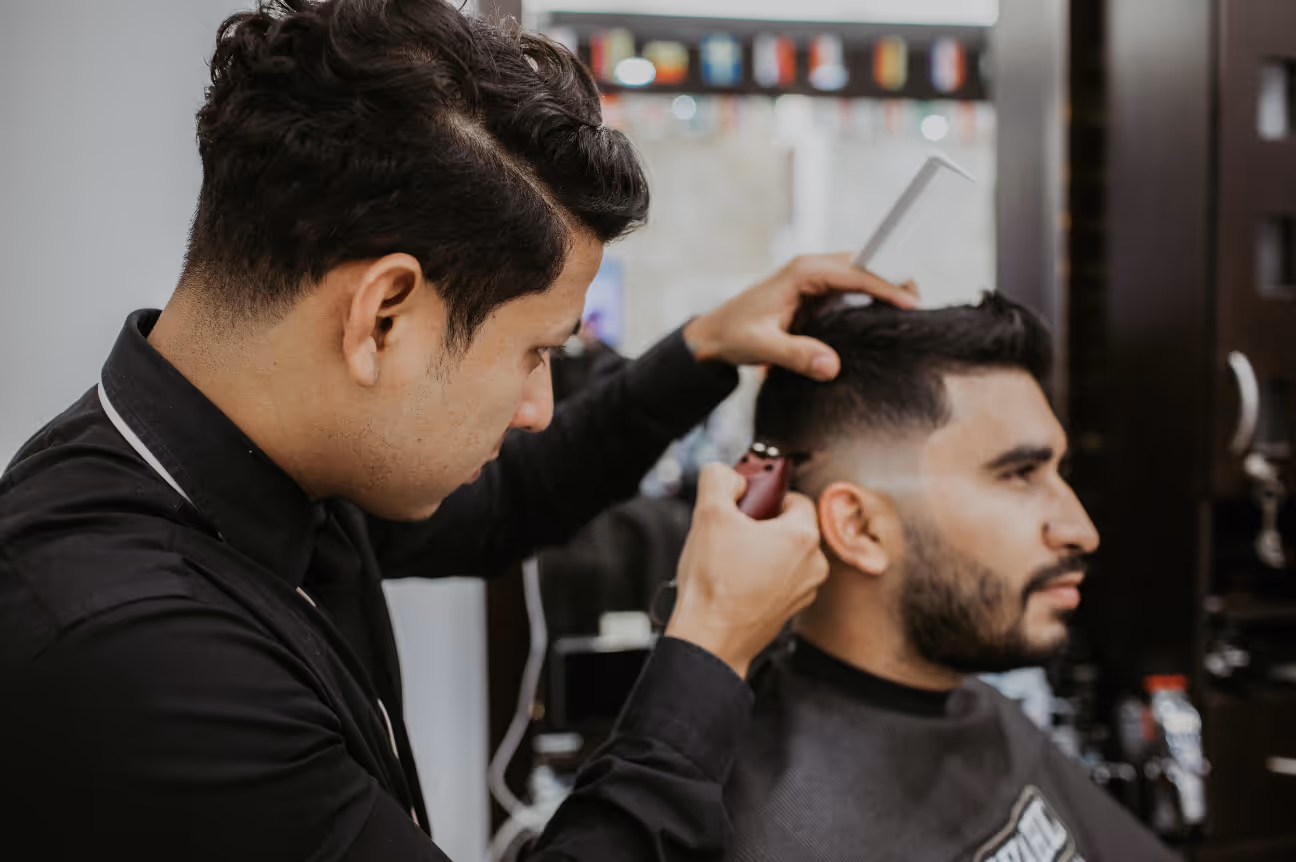 A barber trims a client’s hair with electric clippers while holding a comb, demonstrating typical salon noise exposure.