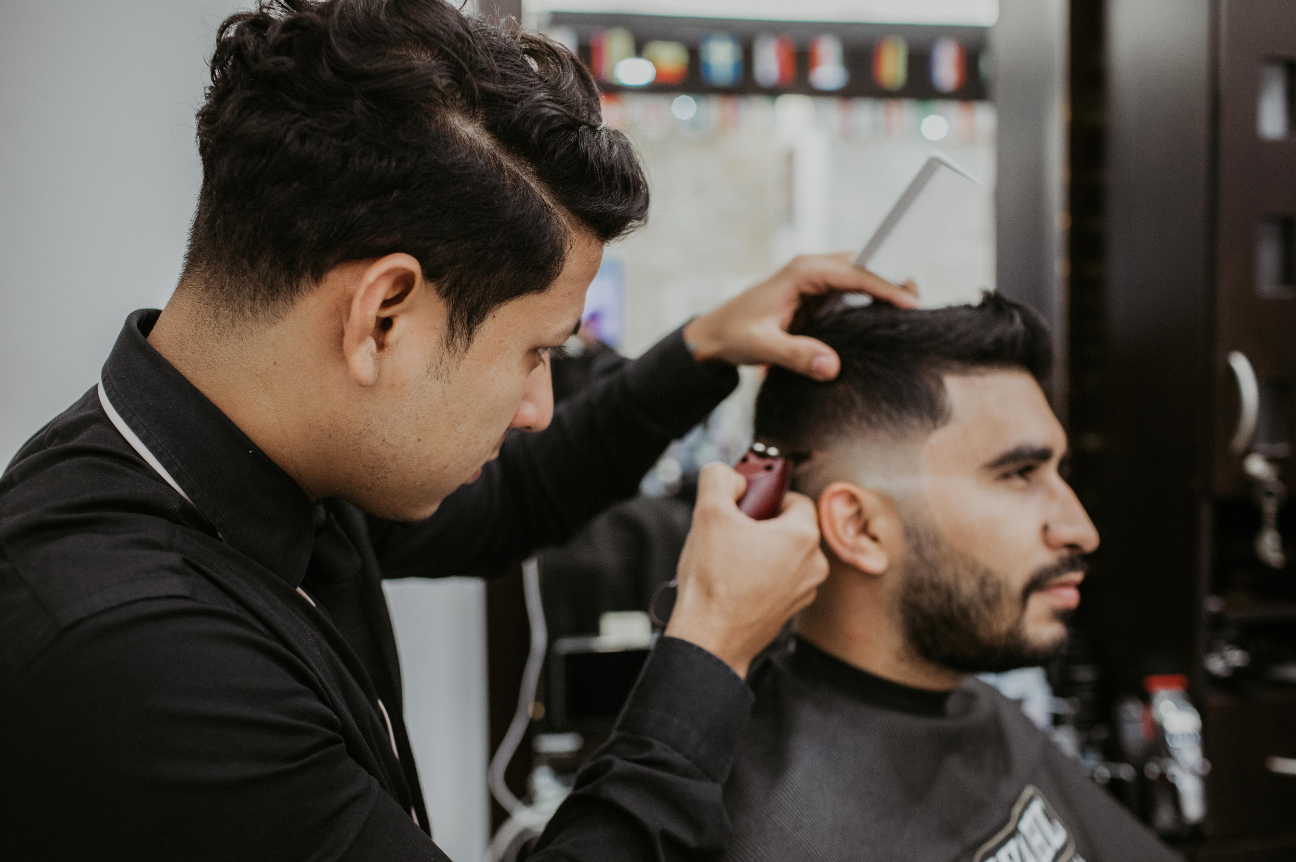 A barber trims a client’s hair with electric clippers while holding a comb, demonstrating typical salon noise exposure.
