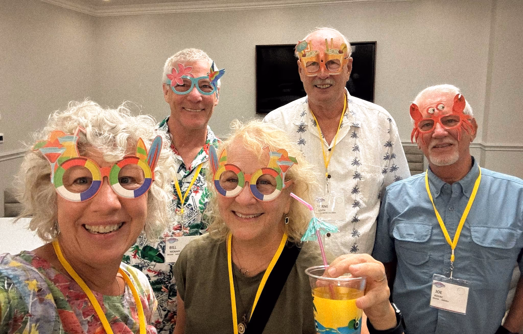 Five SayWhatClub members at the 2023 Galveston convention welcome party, wearing colorful novelty glasses and smiling at the camera, one holding a tropical drink.