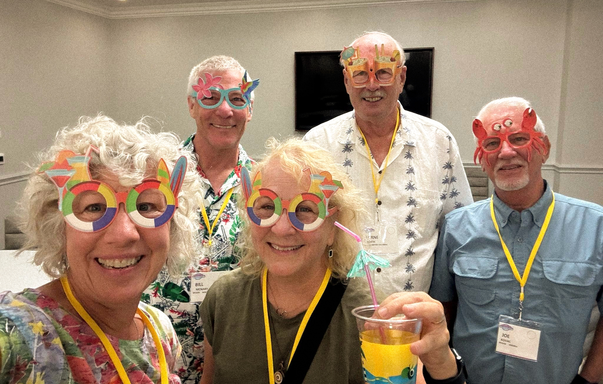 Five SayWhatClub members at the 2023 Galveston convention welcome party, wearing colorful novelty glasses and smiling at the camera, one holding a tropical drink.