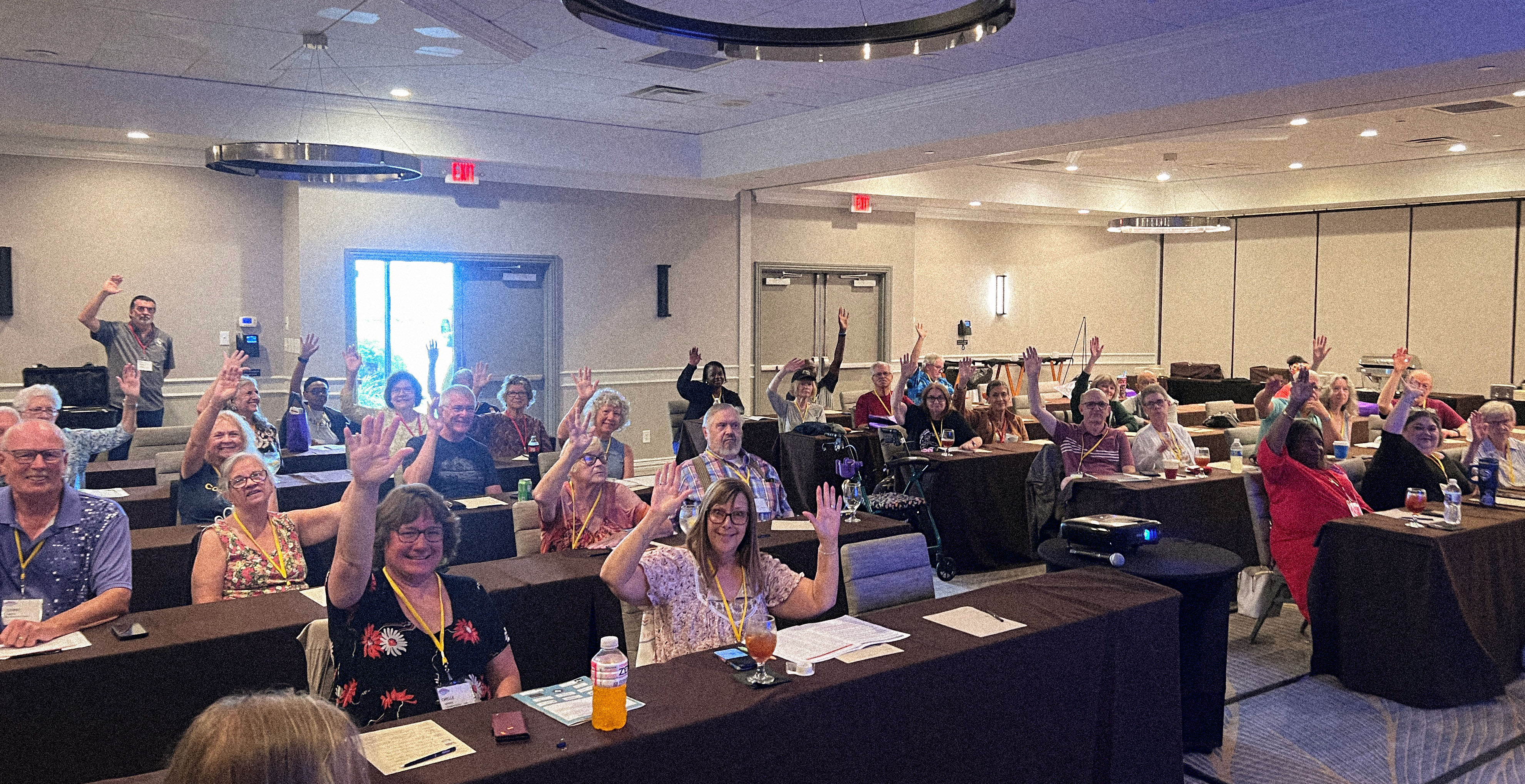 A room full of SayWhatClub members seated at tables during a workshop session, many smiling and raising their hands in participation.