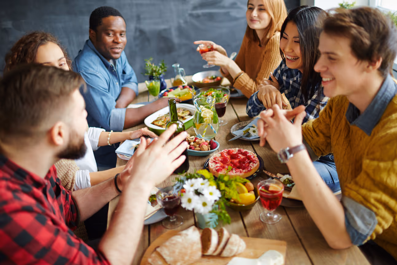 A diverse group of young adults sitting around a dining table, sharing food and conversation. The image illustrates the social situations where people with untreated hearing loss may struggle to follow discussions, highlighting the importance of hearing support for inclusion and connection.