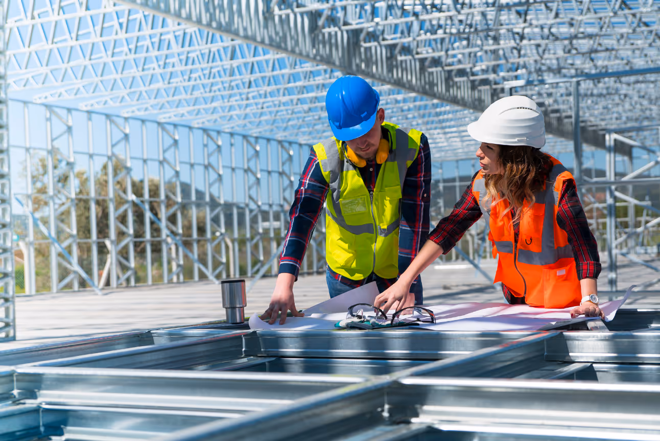 Two construction workers wearing helmets and reflective vests reviewing blueprints at a steel-frame job site. The photo emphasizes the high noise exposure faced by construction professionals and the importance of hearing protection to prevent occupational hearing loss.