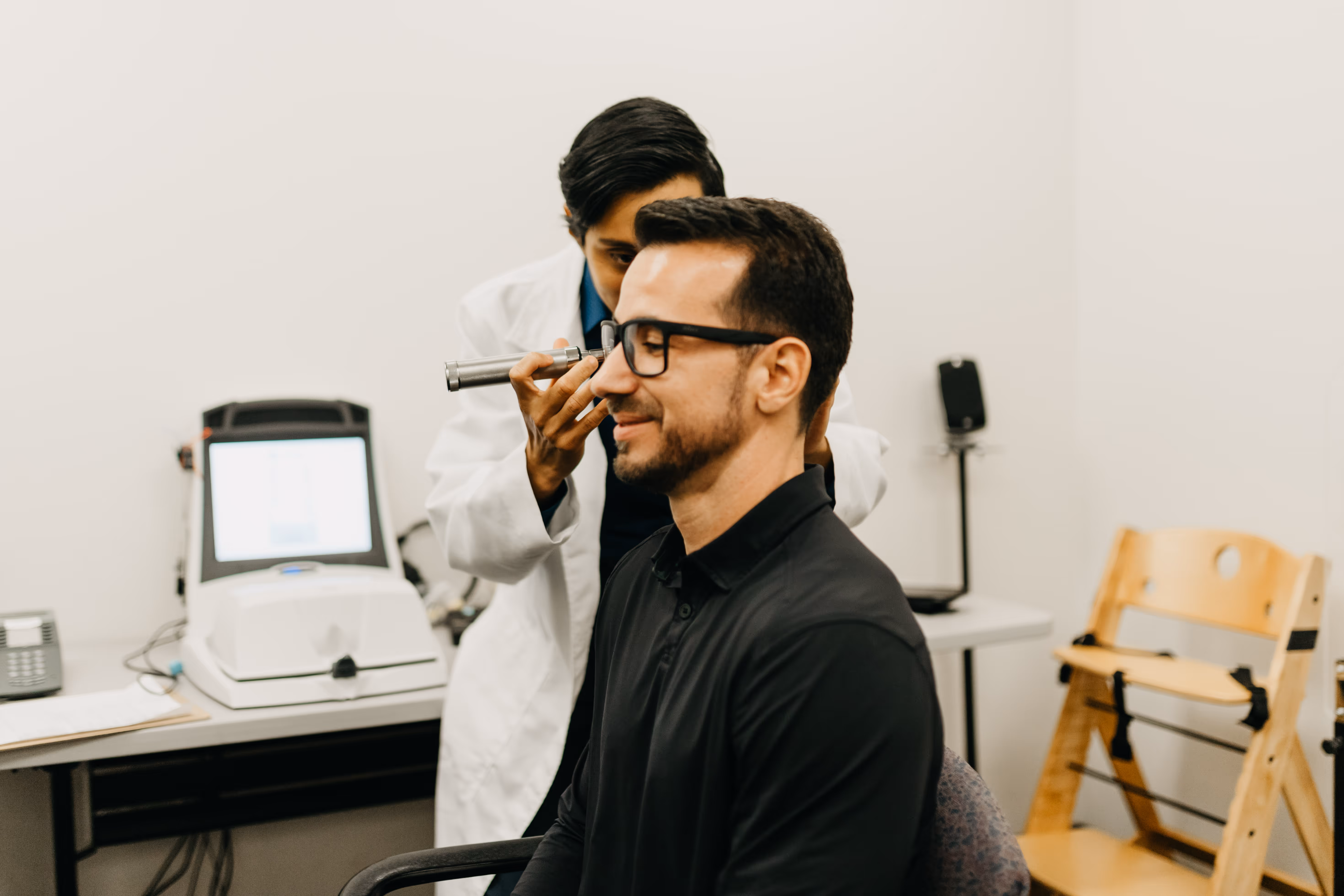 An audiologist in a white coat examines a male patient’s ear with an otoscope during a hearing check-up. The patient, wearing glasses and a black shirt, sits in a testing room equipped with diagnostic machines. The image highlights the importance of routine hearing screenings for early detection of hearing loss.
