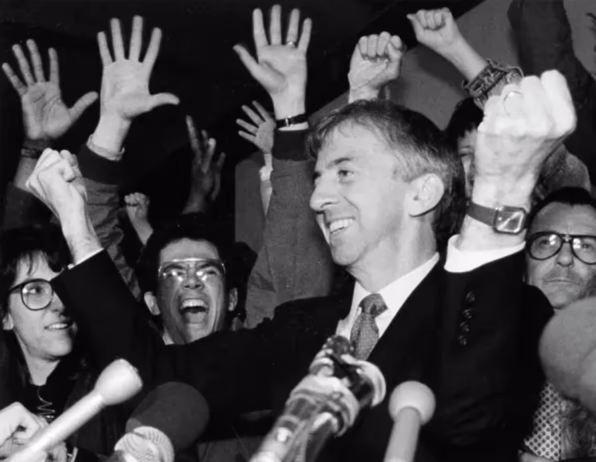 A smiling man in a suit raises his arms in victory while surrounded by cheering supporters with raised hands during a historic announcement. Microphones are visible in the foreground.