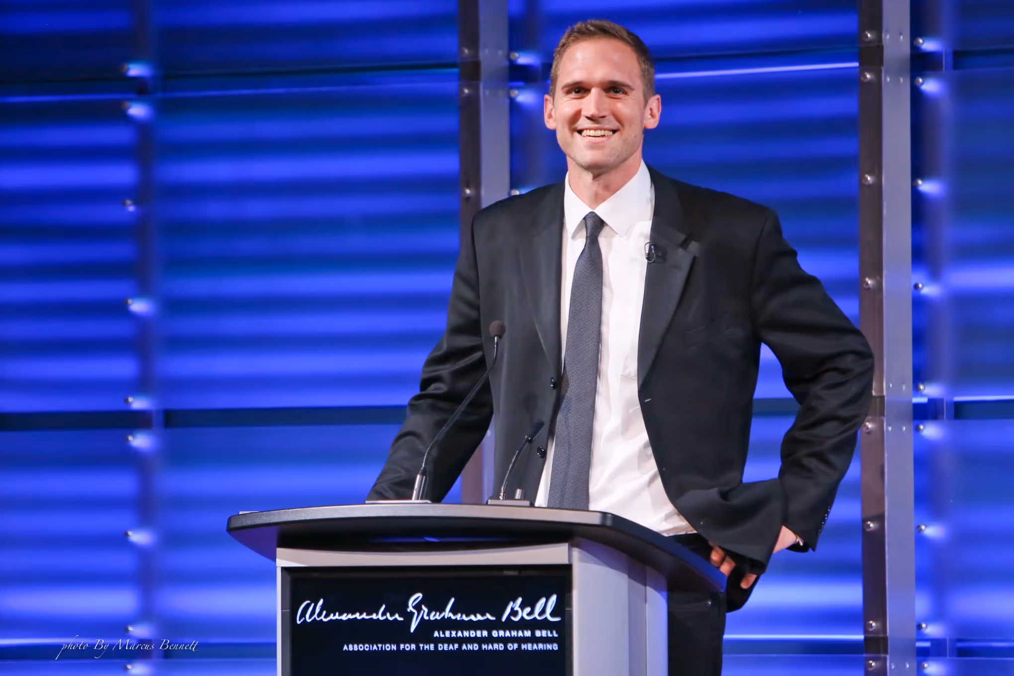 A man in a suit and tie smiles while standing at a podium with microphones, delivering a speech against a blue-lit background.