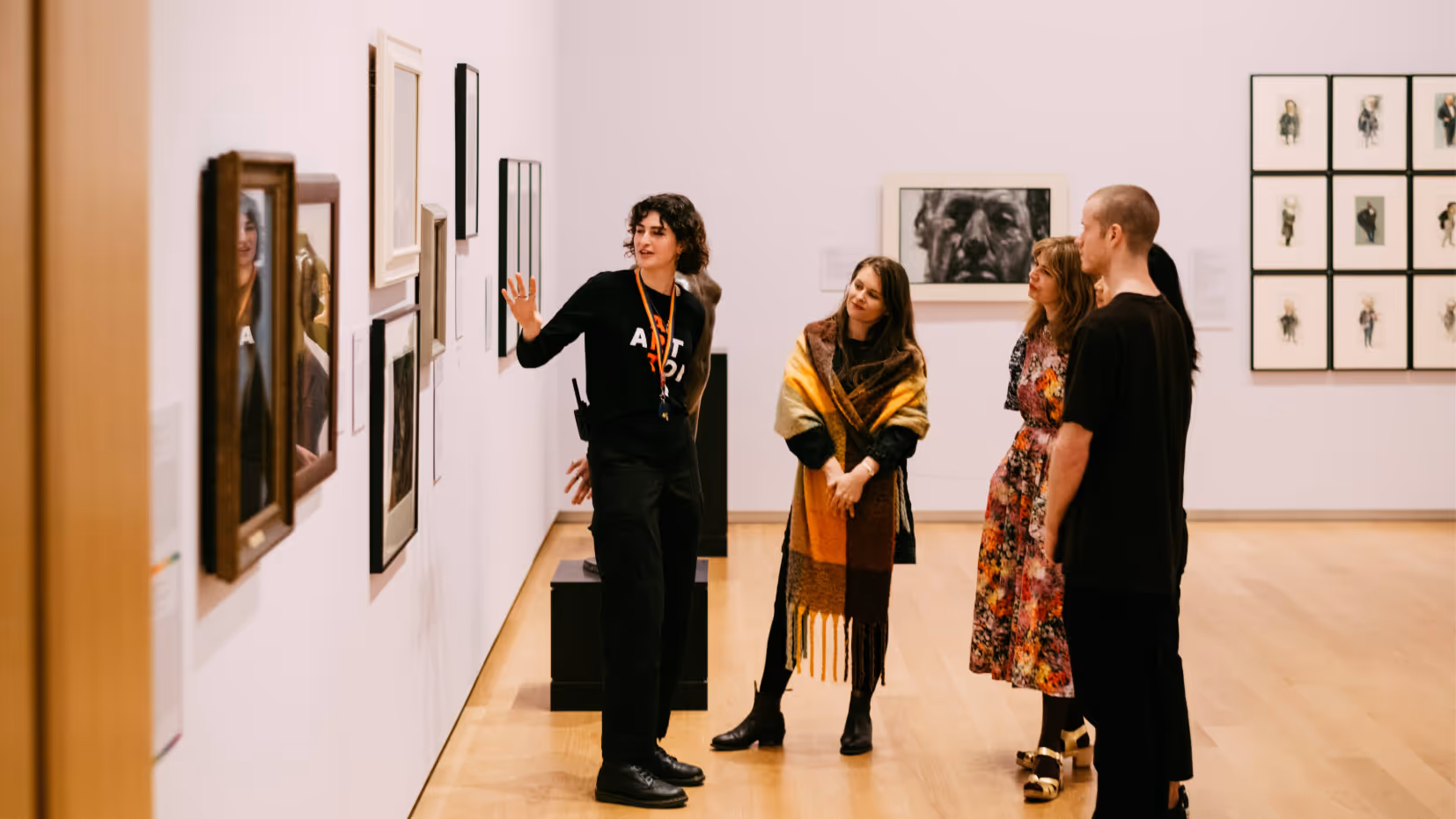 A docent leads a small group through an art gallery, gesturing toward a wall of framed artworks as visitors listen attentively.