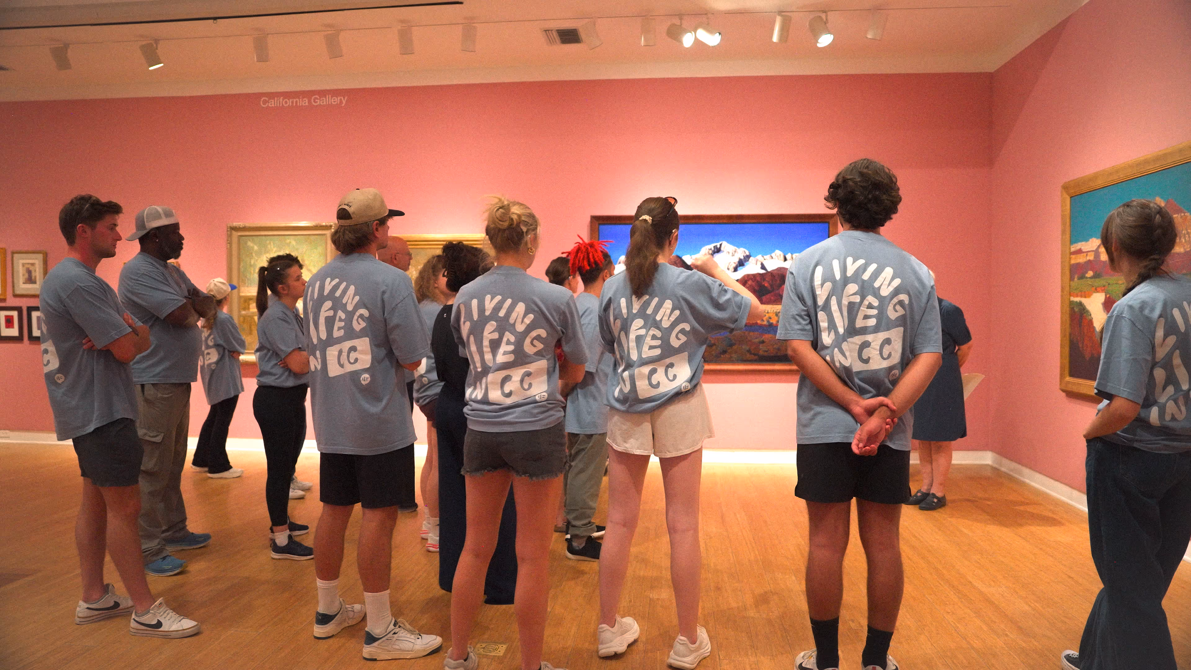 A group of Empower Retreat participants wearing matching blue “Living Life in CC” shirts stands in a gallery at the Laguna Art Museum, listening to a guide while viewing colorful paintings on pink walls.