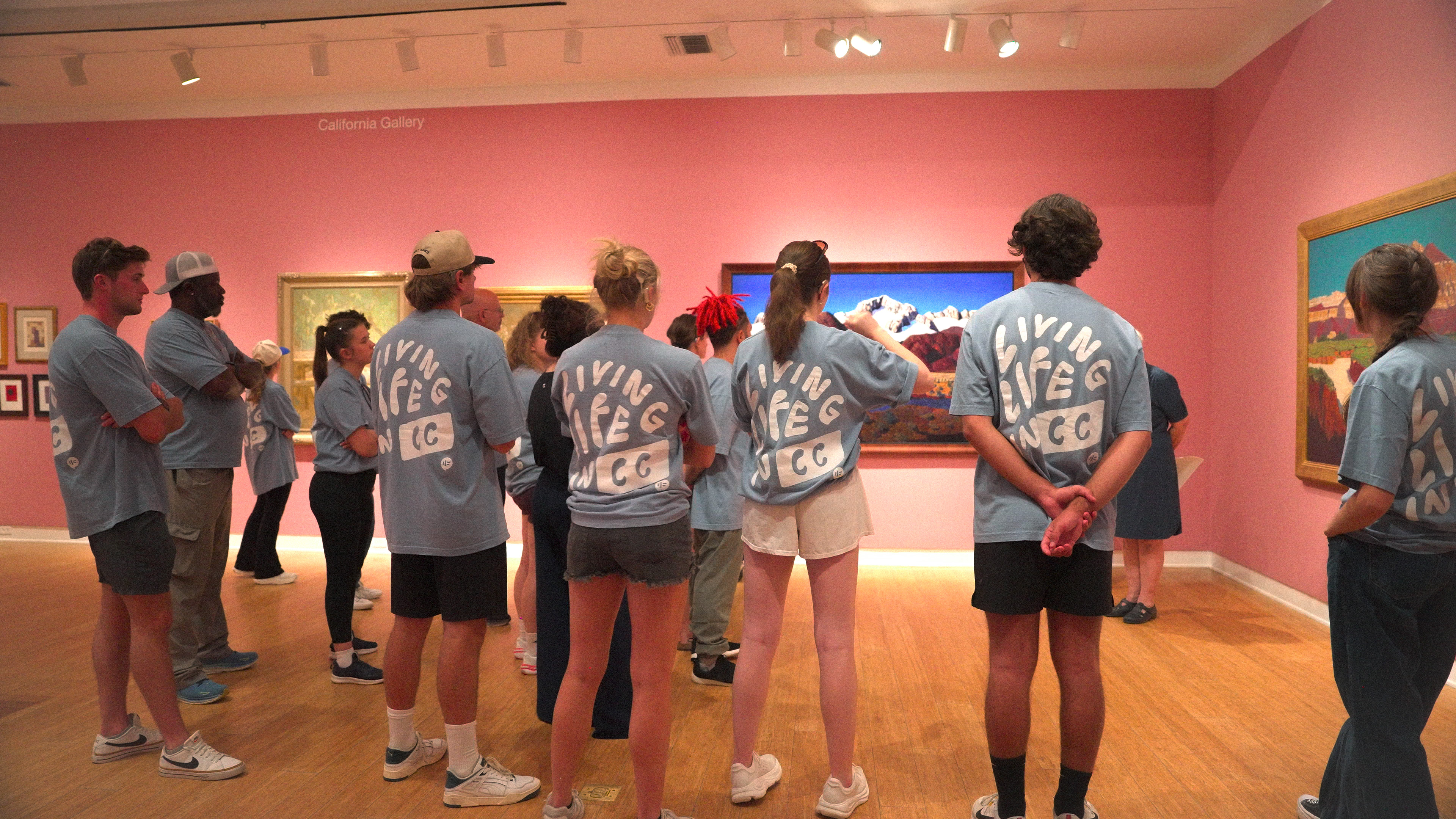 A group of Empower Retreat participants wearing matching blue “Living Life in CC” shirts stands in a gallery at the Laguna Art Museum, listening to a guide while viewing colorful paintings on pink walls.