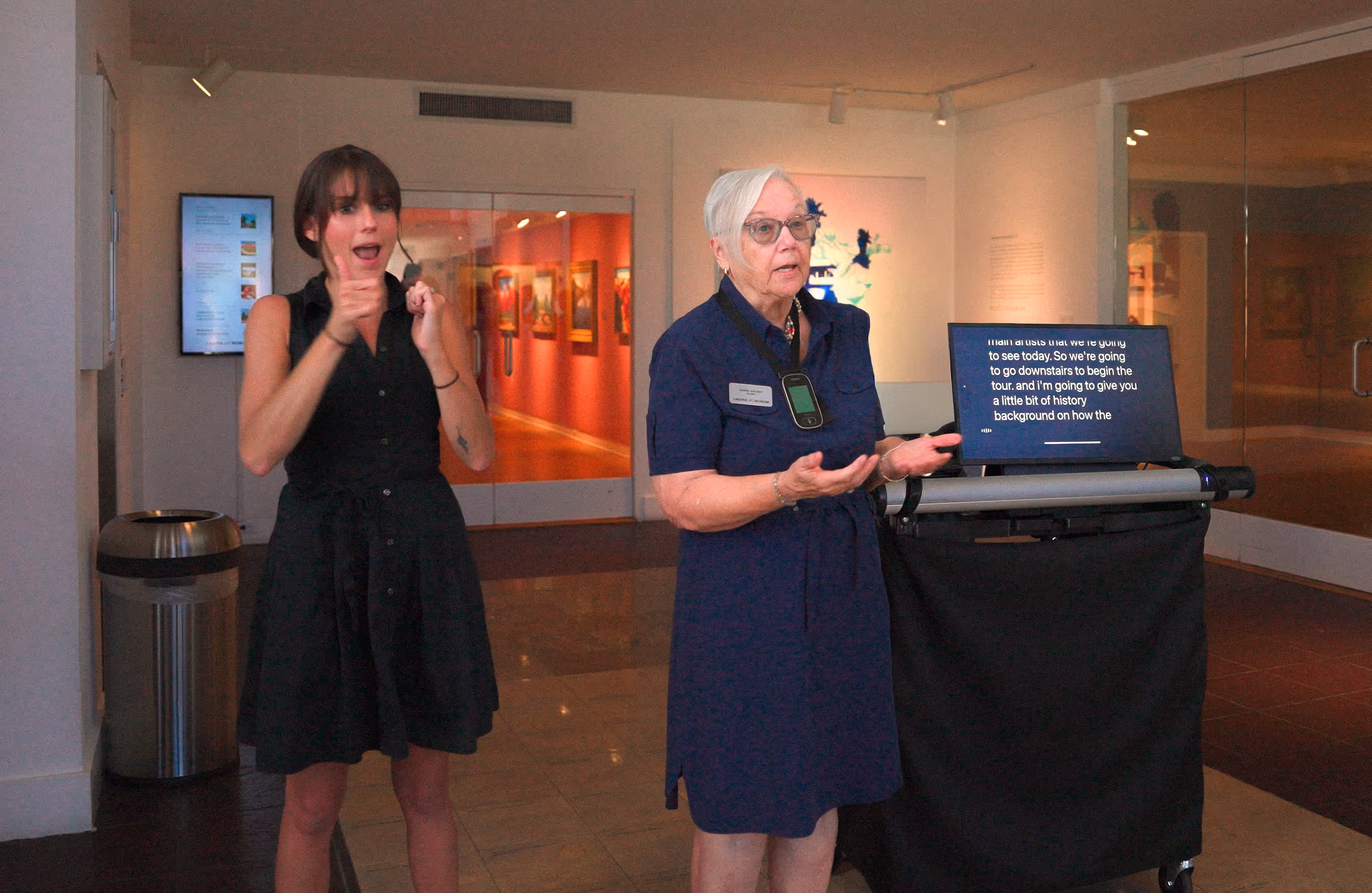A docent speaks beside a mobile captioning cart displaying live captions while an ASL interpreter signs for visitors at the Laguna Art Museum.
