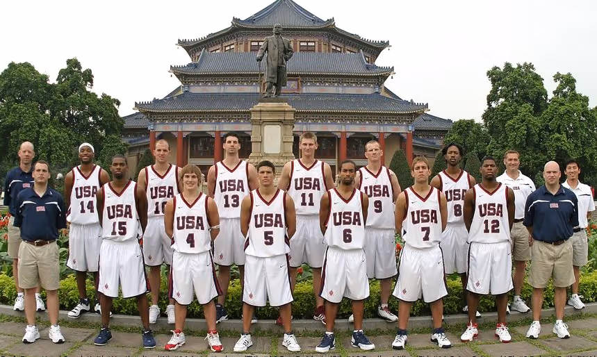 Team USA Deaf Men’s Basketball players pose together in their white uniforms with “USA” printed in red across the chest. The team stands in two rows outside an ornate traditional building with blue-tiled roofs and a bronze statue in front.