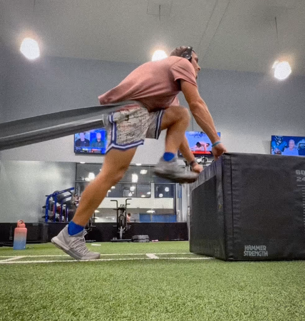 An athlete trains indoors using a resistance band while performing a box jump on turf.