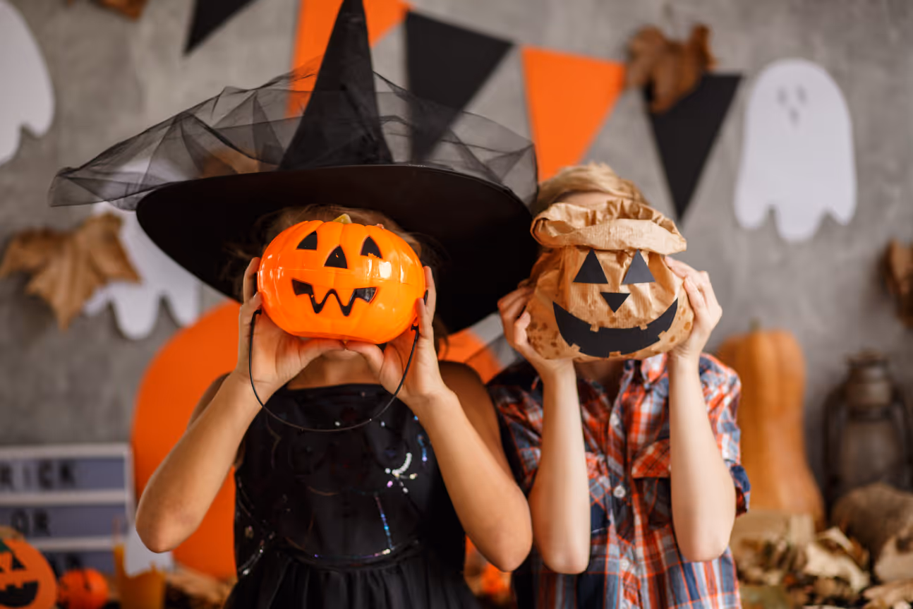 Two children in Halloween costumes playfully hold pumpkin containers over their faces, surrounded by festive orange and black decorations.